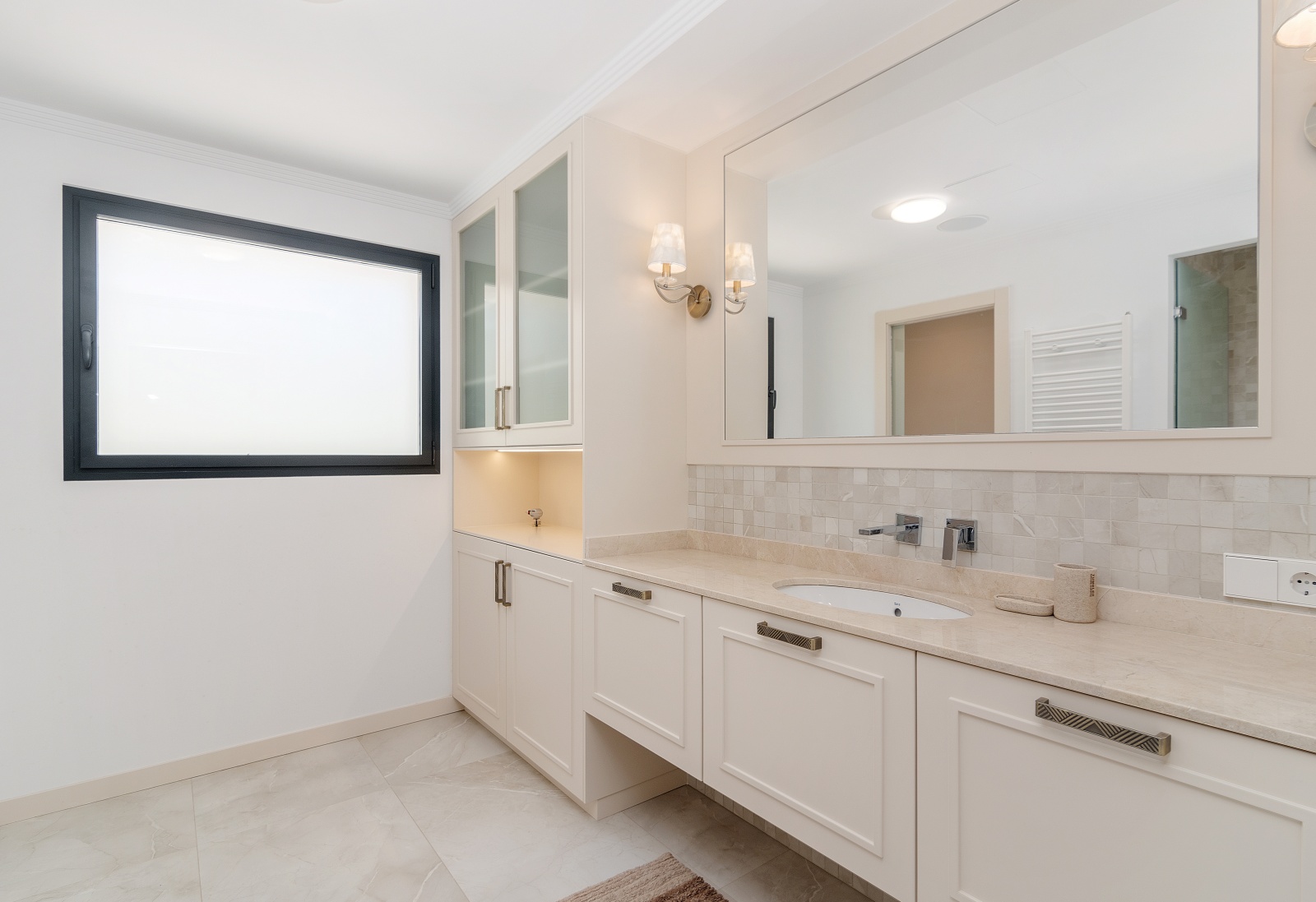 Baño moderno con mueble de lavabo en tono crema, encimera de piedra, armarios empotrados con vitrinas y espejo de gran formato con apliques elegantes.