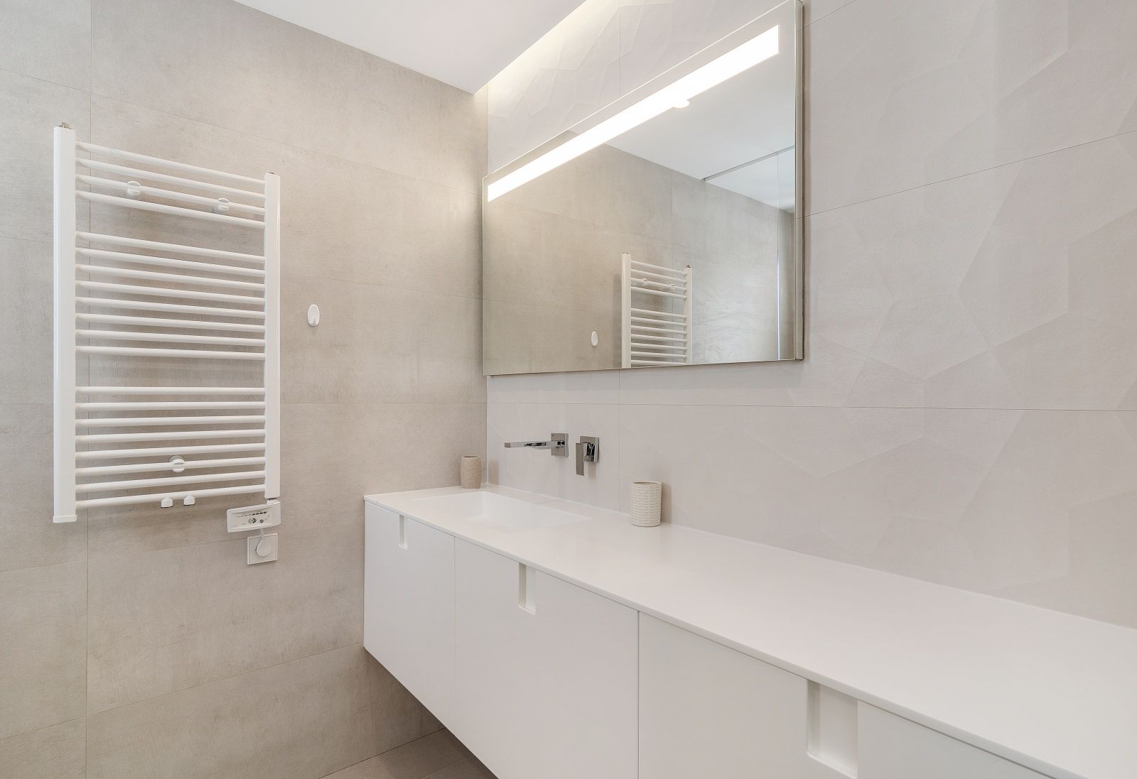 Baño moderno con mueble suspendido blanco, grifería empotrada, espejo con iluminación LED y radiador toallero sobre paredes con relieve geométrico.