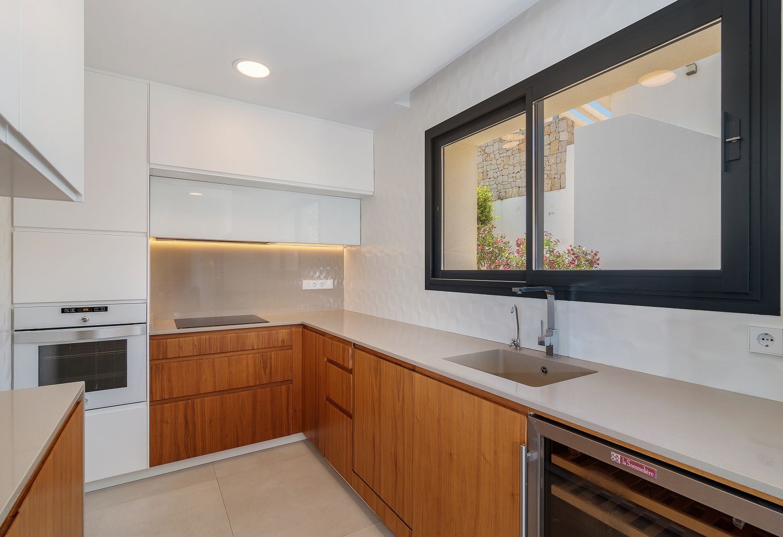 Cocina de diseño con armarios de madera y blanco, encimera de piedra, electrodomésticos integrados, vinoteca y gran ventanal con marco negro hacia el exterior.