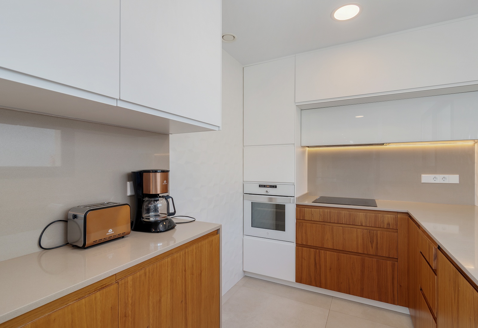 Cocina de diseño con armarios superiores blancos y muebles inferiores de madera, equipada con horno integrado, placa de inducción y encimera de cuarzo.