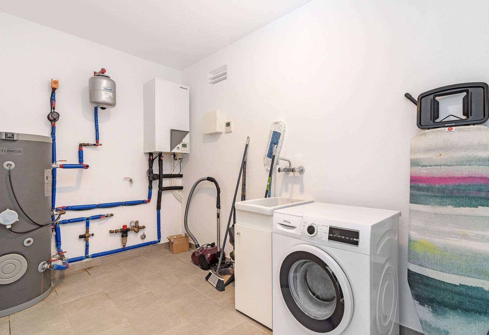 Cuarto técnico y de lavandería equipado con lavadora, fregadero, sistema de aerotermia Daikin y depósito de agua sobre suelo cerámico.
