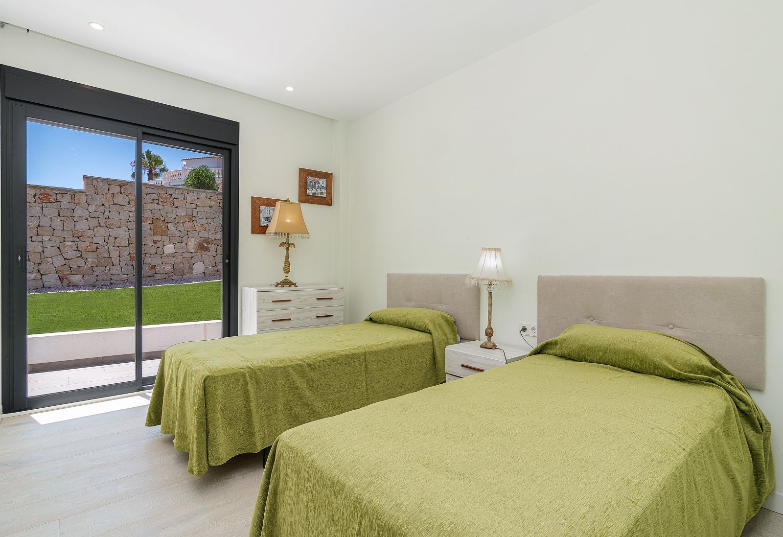 Dormitorio con dos camas individuales, cabeceros tapizados y acceso directo al jardín mediante ventanal corredero. Suelo de gres porcelánico imitación madera y paredes blancas.
