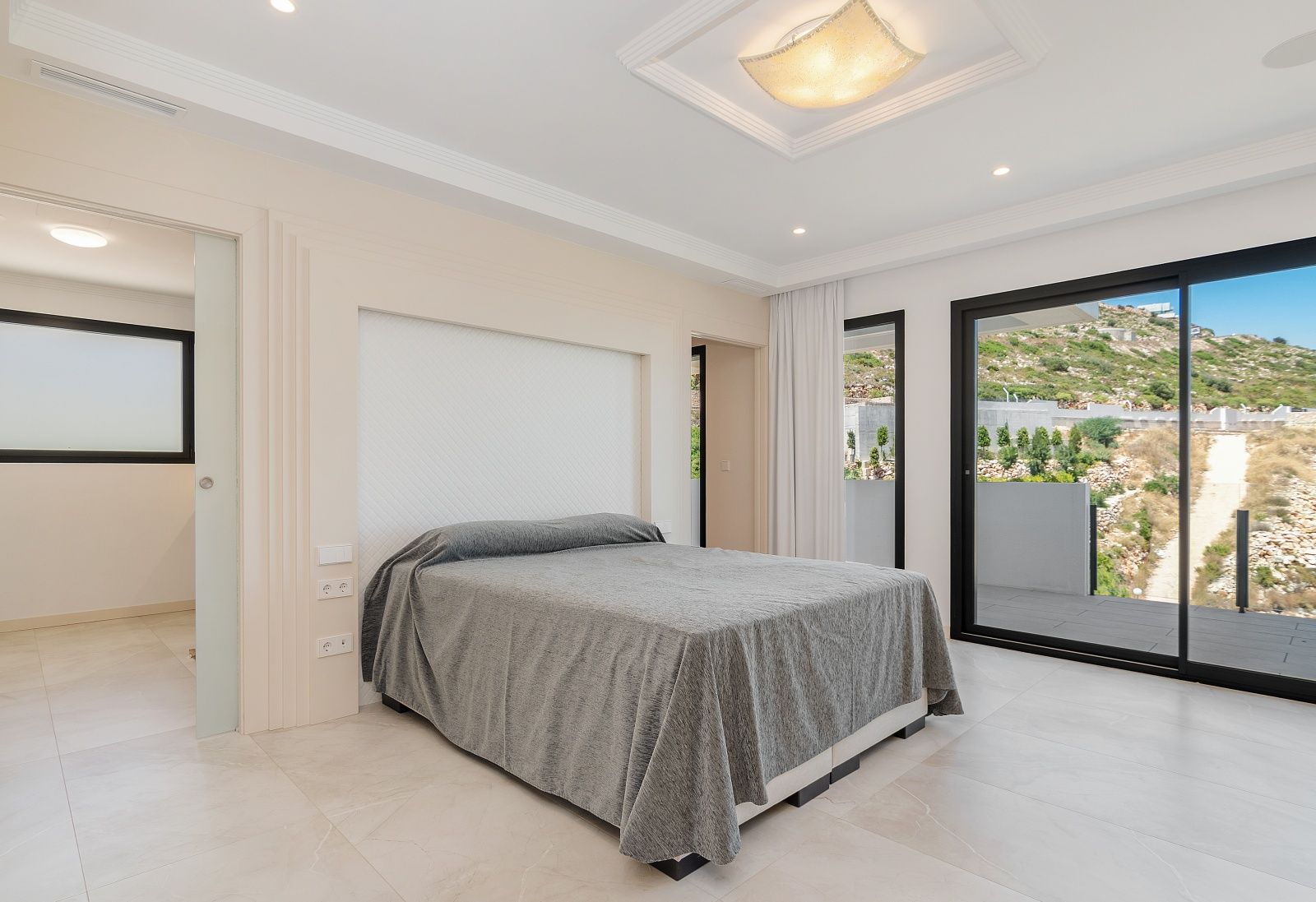 Dormitorio luminoso con acceso directo a la terraza, suelos cerámicos, iluminación empotrada y cabecero integrado con molduras decorativas.