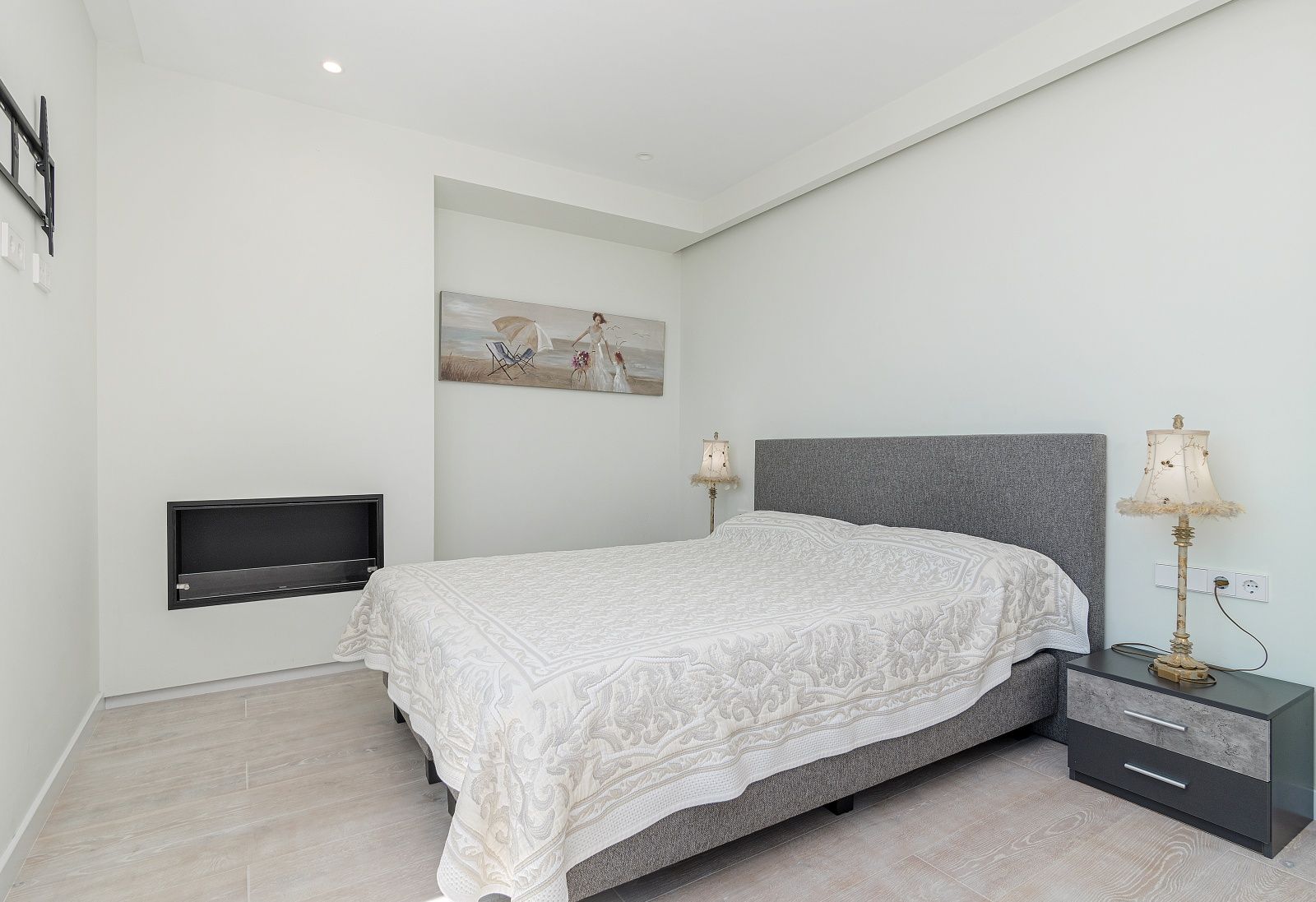Dormitorio moderno con chimenea encastrada, cama con cabecero gris, mesita de noche de diseño y suelos de gres porcelánico imitación madera clara.
