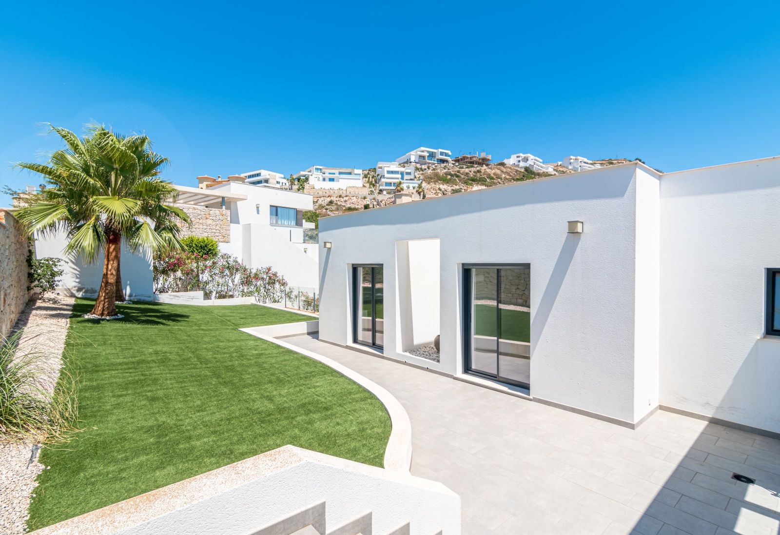 Fachada blanca de diseño moderno con grandes ventanales, zona de césped artificial y palmera, integrada en el entorno residencial de Cumbre del Sol.