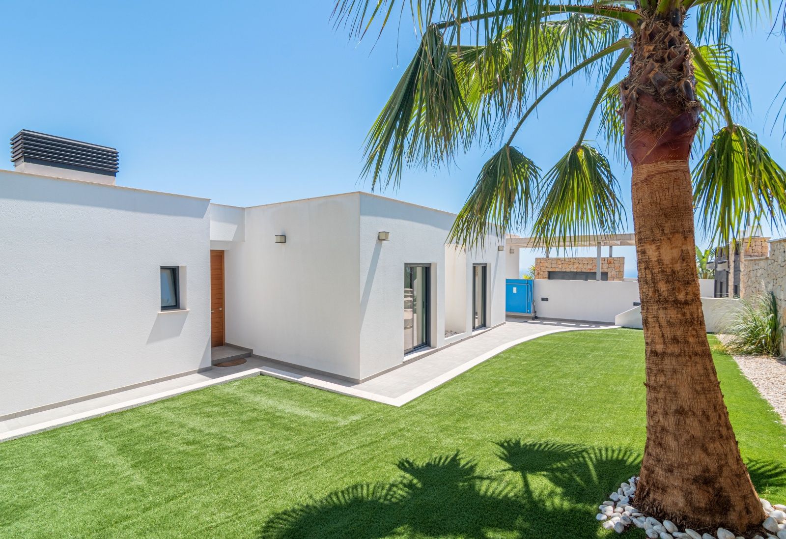 Fachada blanca de estilo moderno con jardín de césped artificial, palmera perimetral y acceso principal en madera bajo un cielo despejado.