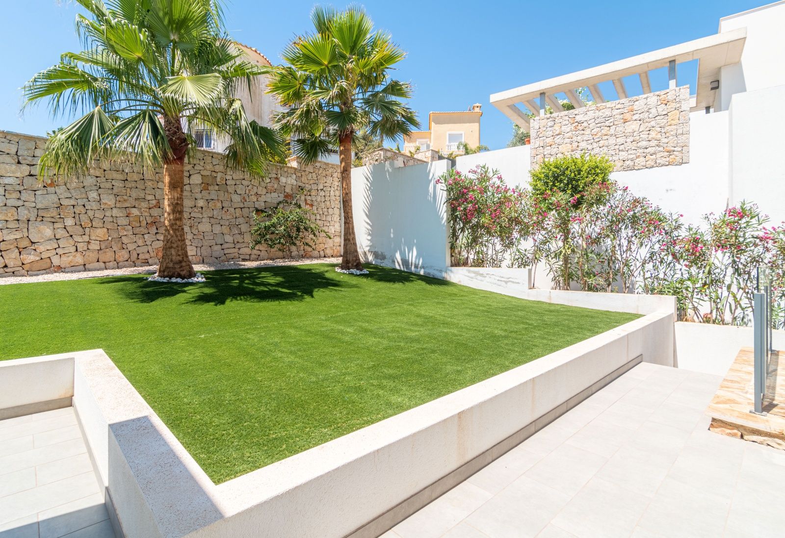Jardín de diseño de bajo mantenimiento con césped artificial, palmeras y muros de piedra natural integrados en la arquitectura moderna de la villa.