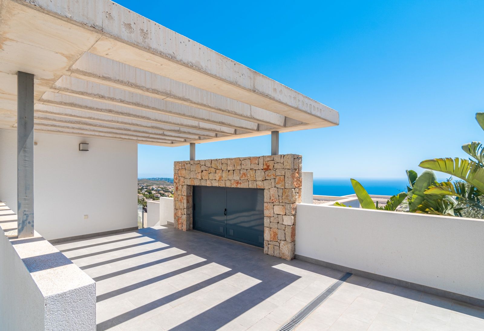 Parking techado con pérgola de hormigón visto y muro de piedra natural, ofreciendo vistas panorámicas al mar Mediterráneo y un diseño arquitectónico moderno.