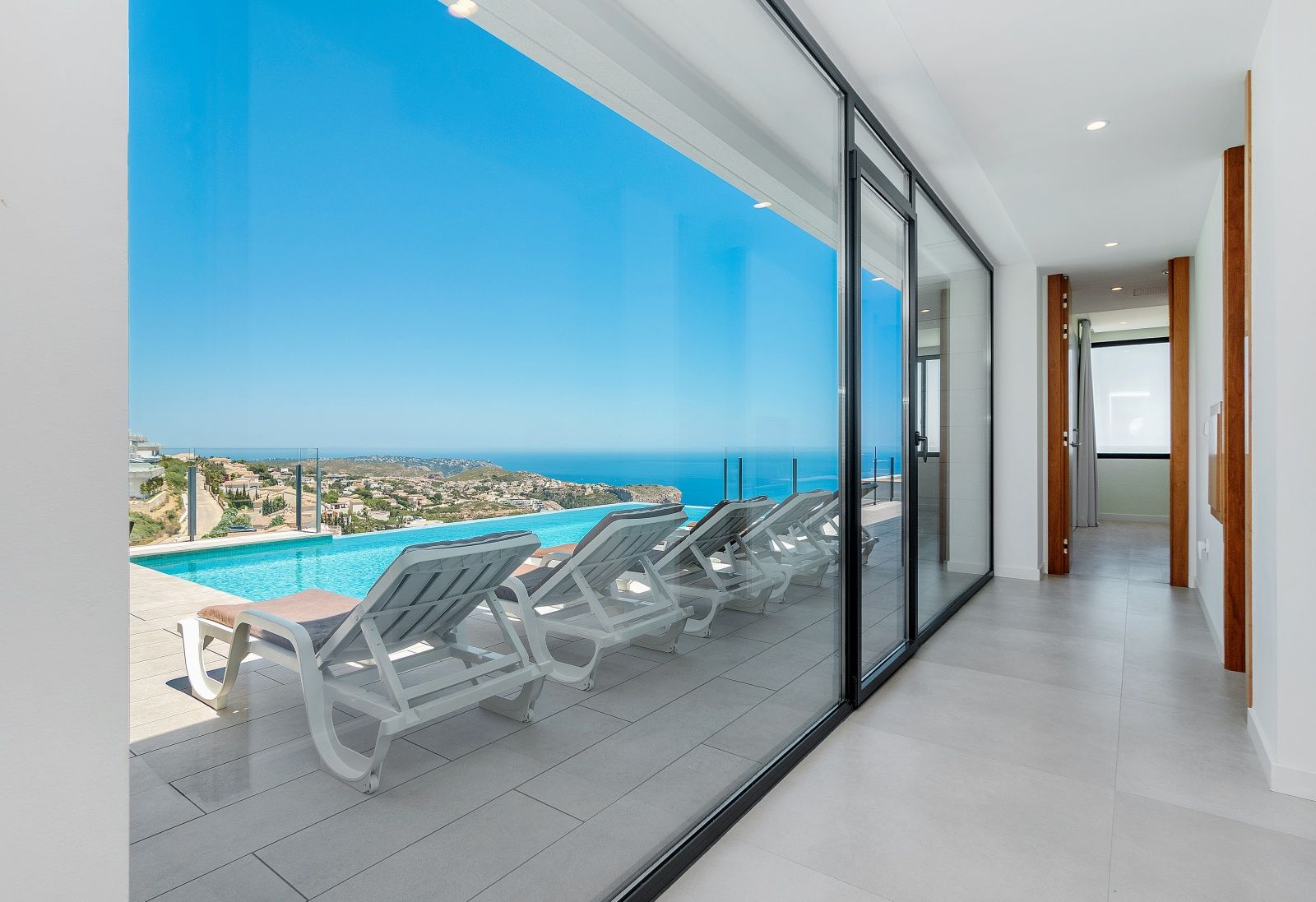 Pasillo interior con grandes ventanales correderos que ofrecen acceso directo a la terraza de la piscina infinita y vistas panorámicas al mar Mediterráneo.