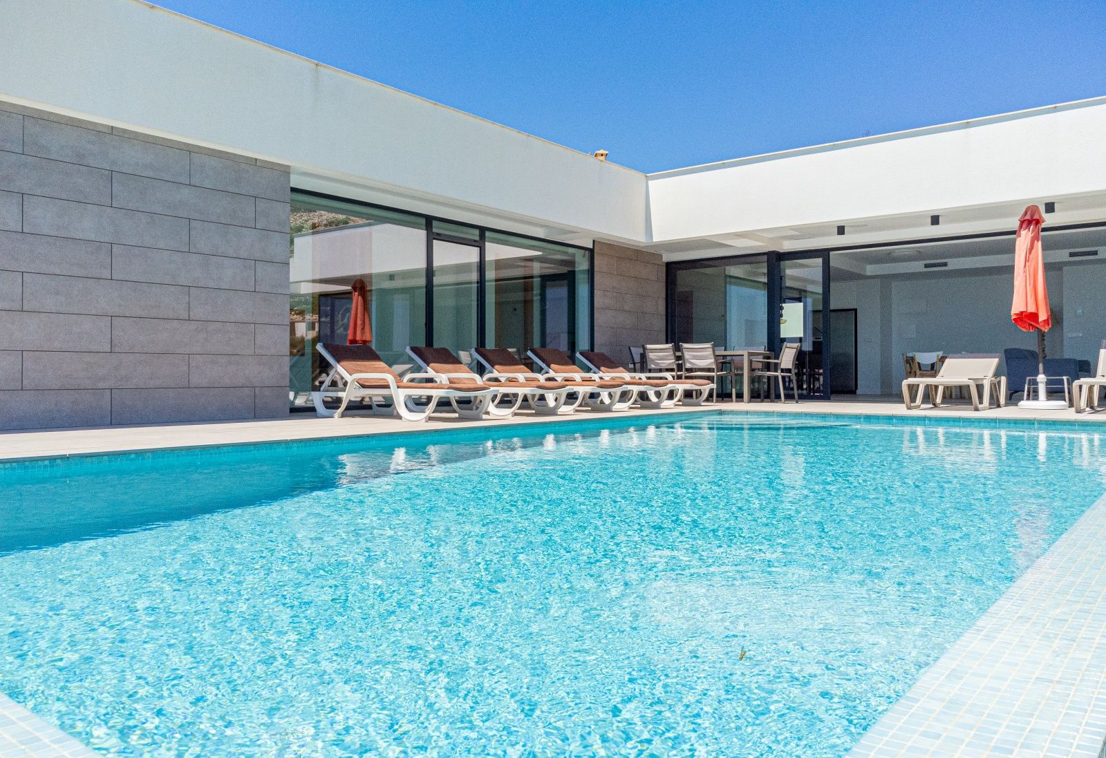 Piscina rectangular con gresite azul y amplia terraza solárium. La fachada moderna combina paneles de piedra gris con grandes ventanales y una zona de comedor exterior.