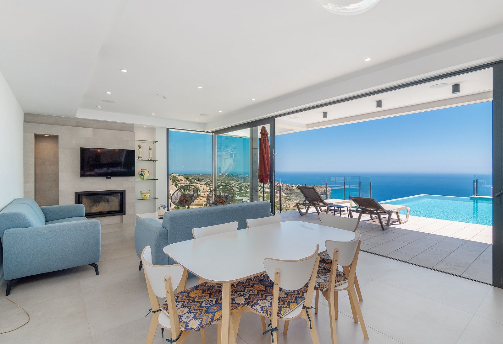 Salón comedor de concepto abierto con chimenea y amplios ventanales que integran el interior con la terraza, la piscina infinita y las vistas al mar.