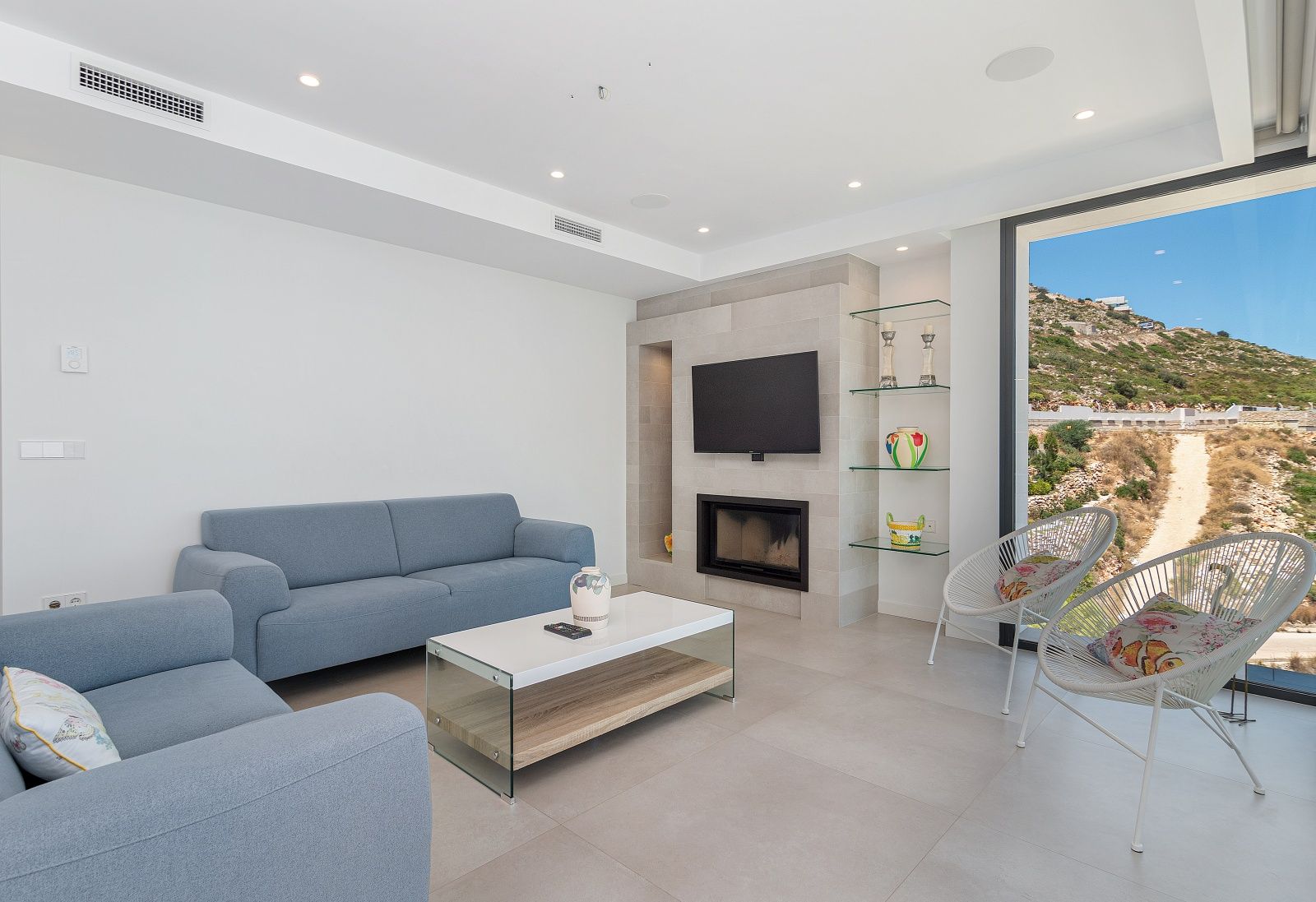 Salón contemporáneo con sofás azules, chimenea integrada en pared de piedra natural, estanterías de cristal y grandes ventanales con vistas al entorno.