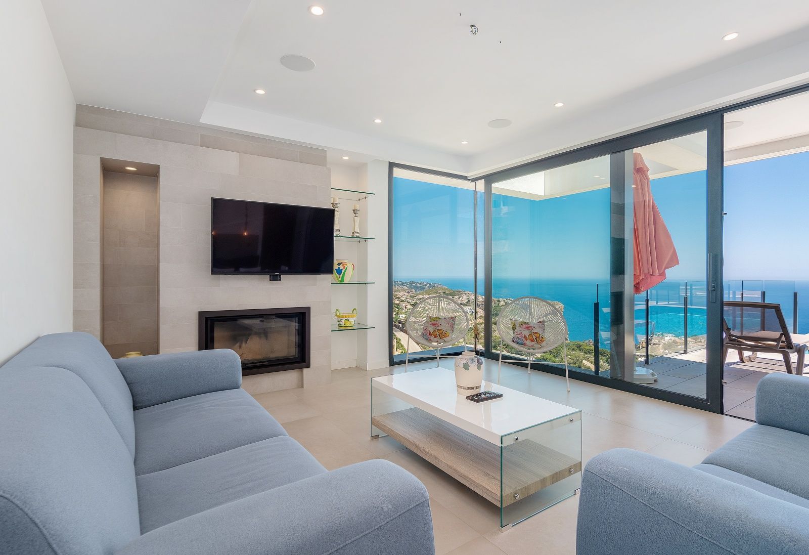 Salón moderno con sofás azules, chimenea integrada y amplios ventanales que ofrecen vistas panorámicas al mar Mediterráneo y la costa de Benitachell.