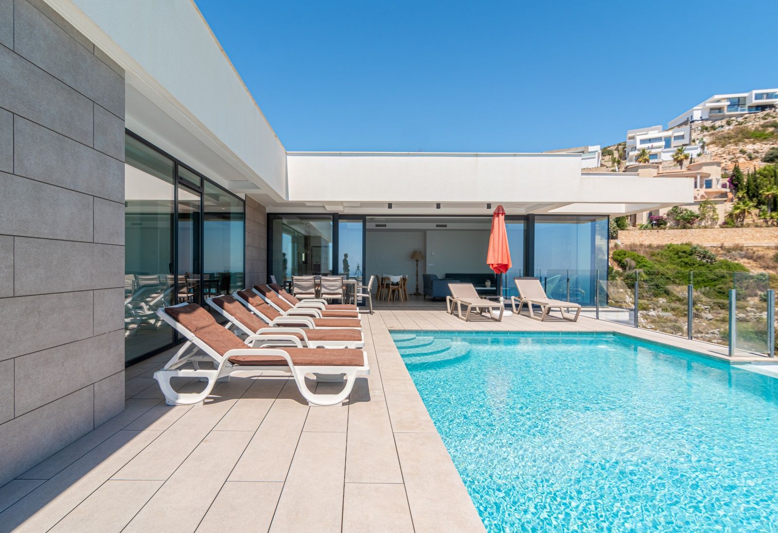 Terraza exterior con piscina infinita, hamacas y amplios ventanales que integran el salón con el área de solárium bajo una arquitectura moderna de líneas blancas.