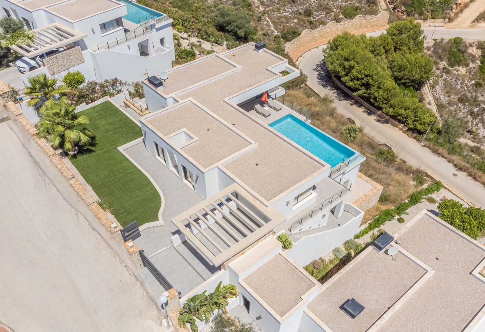 Vista aérea de la villa moderna con piscina infinita, cubiertas de grava, zona de césped artificial y parking con pérgola en Cumbre del Sol.