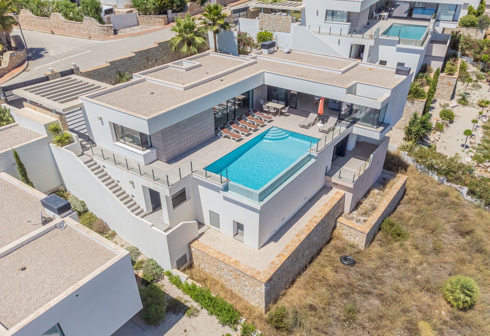 Vista aérea de la villa moderna con piscina infinita de borde de cristal, amplia terraza con tumbonas y zona de comedor exterior bajo una estructura blanca minimalista.