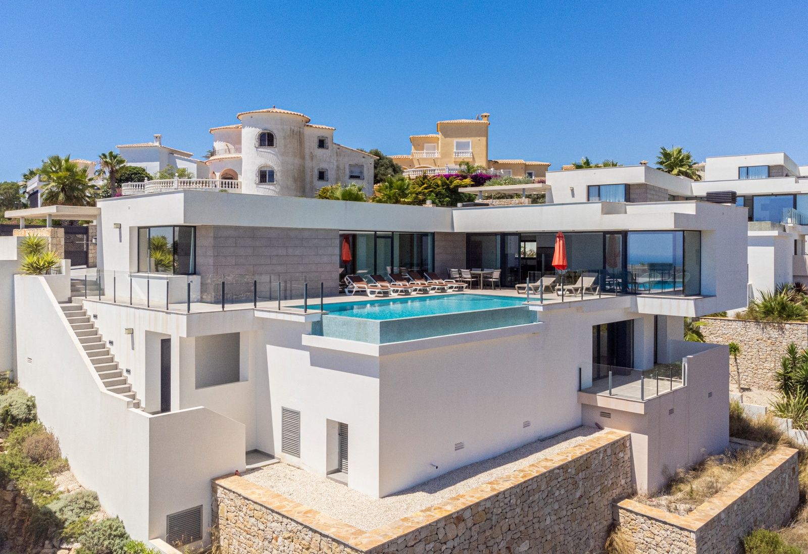 Vista exterior de la villa moderna con piscina infinita, amplias terrazas con barandillas de cristal y fachada blanca con detalles en piedra natural.