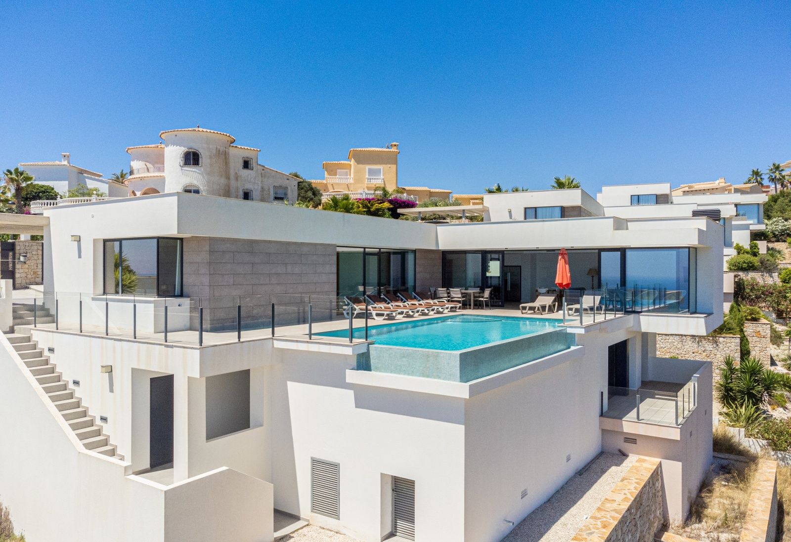 Vista exterior de villa moderna con piscina infinita, terraza con barandillas de cristal y fachada de líneas rectas con detalles en piedra natural.