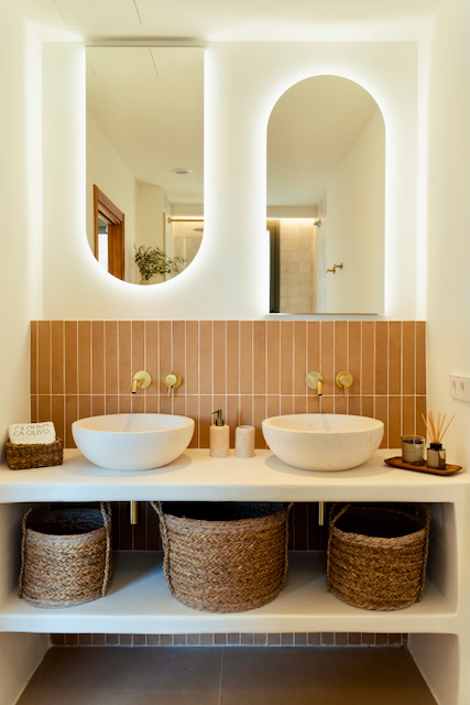 Baño con lavabo doble de piedra, grifería empotrada dorada y azulejos verticales terracota. Incluye espejos arqueados retroiluminados y cestas de mimbre para almacenamiento.