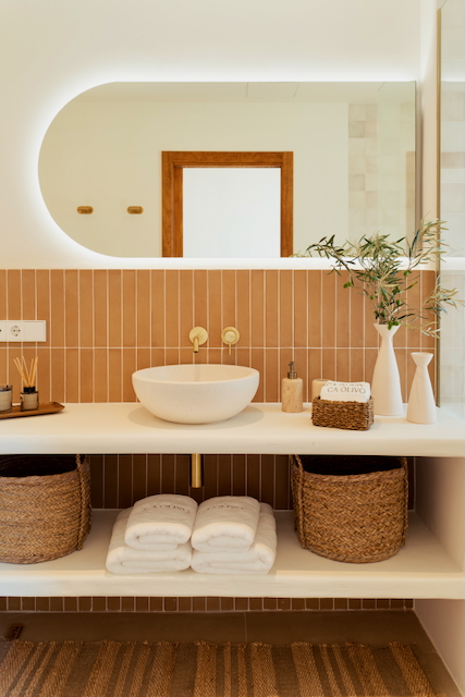 Baño moderno con azulejos verticales en tono terracota, lavabo sobre encimera blanca y grifería en latón dorado. Incluye un espejo retroiluminado y estantes con cestas de mimbre.