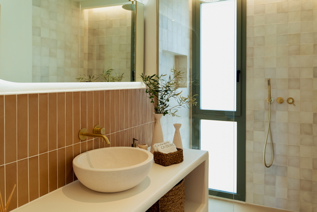 Baño moderno con lavabo de piedra, grifería en oro cepillado y azulejos verticales en tonos tierra. Incluye zona de ducha con mampara de cristal y abundante luz natural.