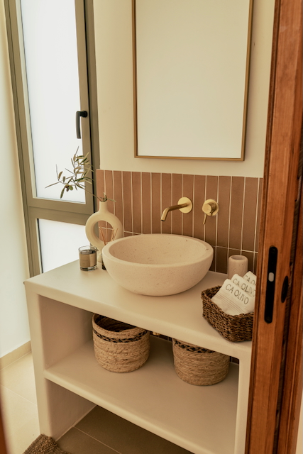 Baño moderno con lavabo de piedra sobre encimera blanca, grifería empotrada en dorado y frente de azulejos verticales en tonos tierra.