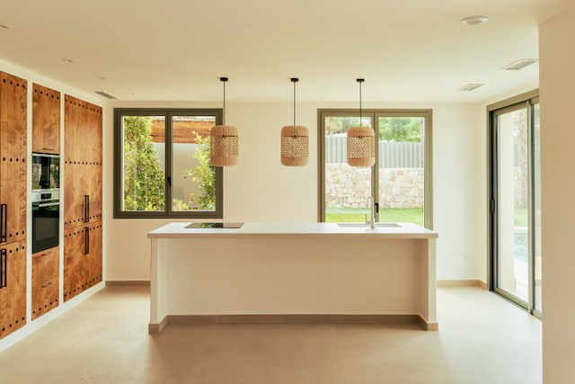 Cocina de concepto abierto con isla blanca, lámparas de fibras naturales y armarios de madera. Los ventanales integran el interior con el jardín y la zona de piscina.