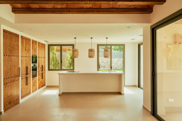 Cocina de diseño abierto con isla blanca, armarios de madera rústica y vigas vistas. Destacan las lámparas de mimbre y el acceso directo al exterior mediante grandes ventanales.