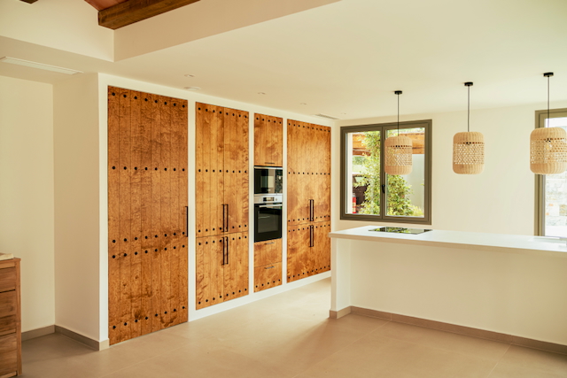 Cocina de diseño con armarios de madera rústica y electrodomésticos integrados. Incluye una isla blanca con placa de inducción y lámparas colgantes de fibras naturales.