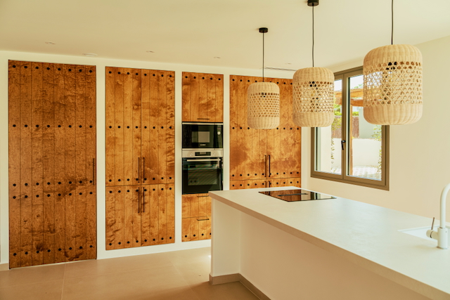 Cocina moderna con isla blanca, electrodomésticos integrados y armarios de madera con detalles de tachuelas. Lámparas de mimbre cuelgan sobre la encimera bajo luz natural.