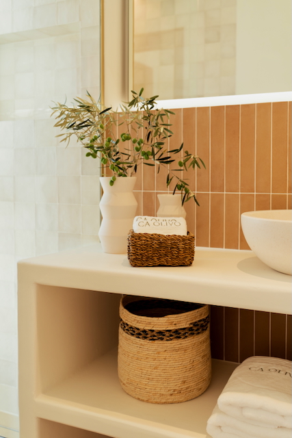 Detalle de baño con encimera de obra, lavabo sobre encimera y revestimiento de azulejos verticales en tonos tierra. Incluye estantes abiertos con cestas de mimbre y decoración natural.