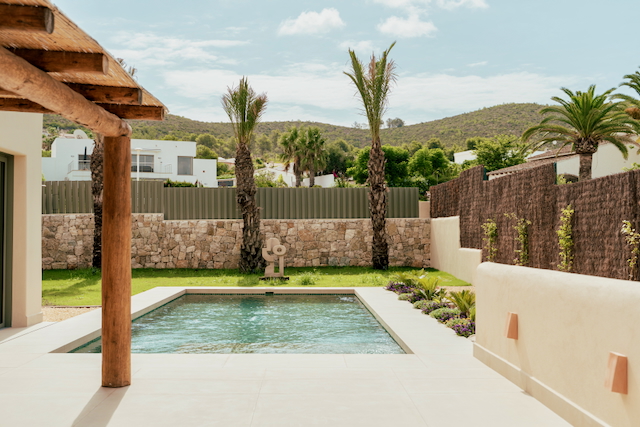 Piscina rectangular integrada en terraza de gres porcelánico, rodeada de muros de piedra natural, jardín mediterráneo con palmeras y porche de vigas de madera rústica.