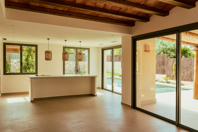 Salón diáfano con vigas de madera vistas, cocina con isla minimalista y lámparas de mimbre, con acceso directo a la piscina mediante amplios ventanales.