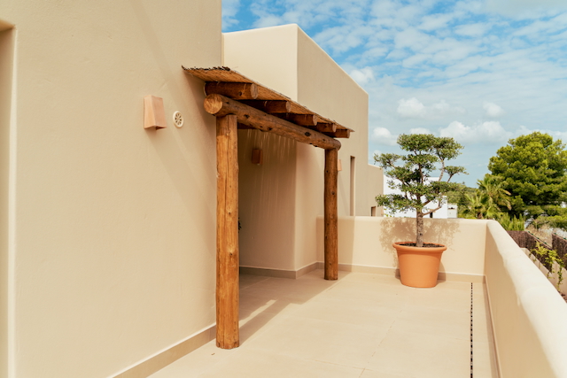 Terraza exterior con pérgola de madera rústica, techado de cañizo y suelos de gres en tonos neutros junto a un árbol ornamental en maceta.