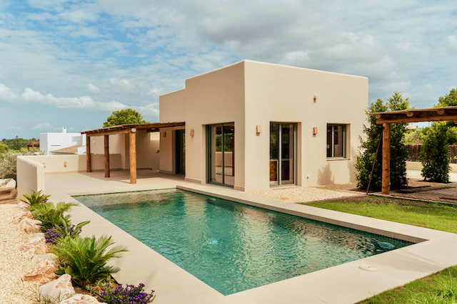 Vista exterior de la villa moderna con piscina rectangular, fachada en tonos arena y una pérgola de madera rústica integrada en la amplia terraza.