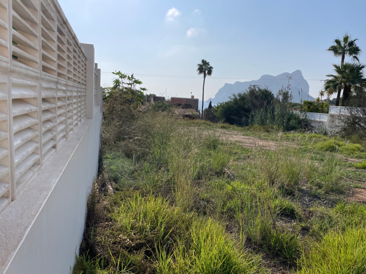Parcela de 856 m² en Bassetes, Calpe, delimitada por un muro blanco con celosía y vistas directas al Peñón de Ifach bajo el sol mediterráneo.
