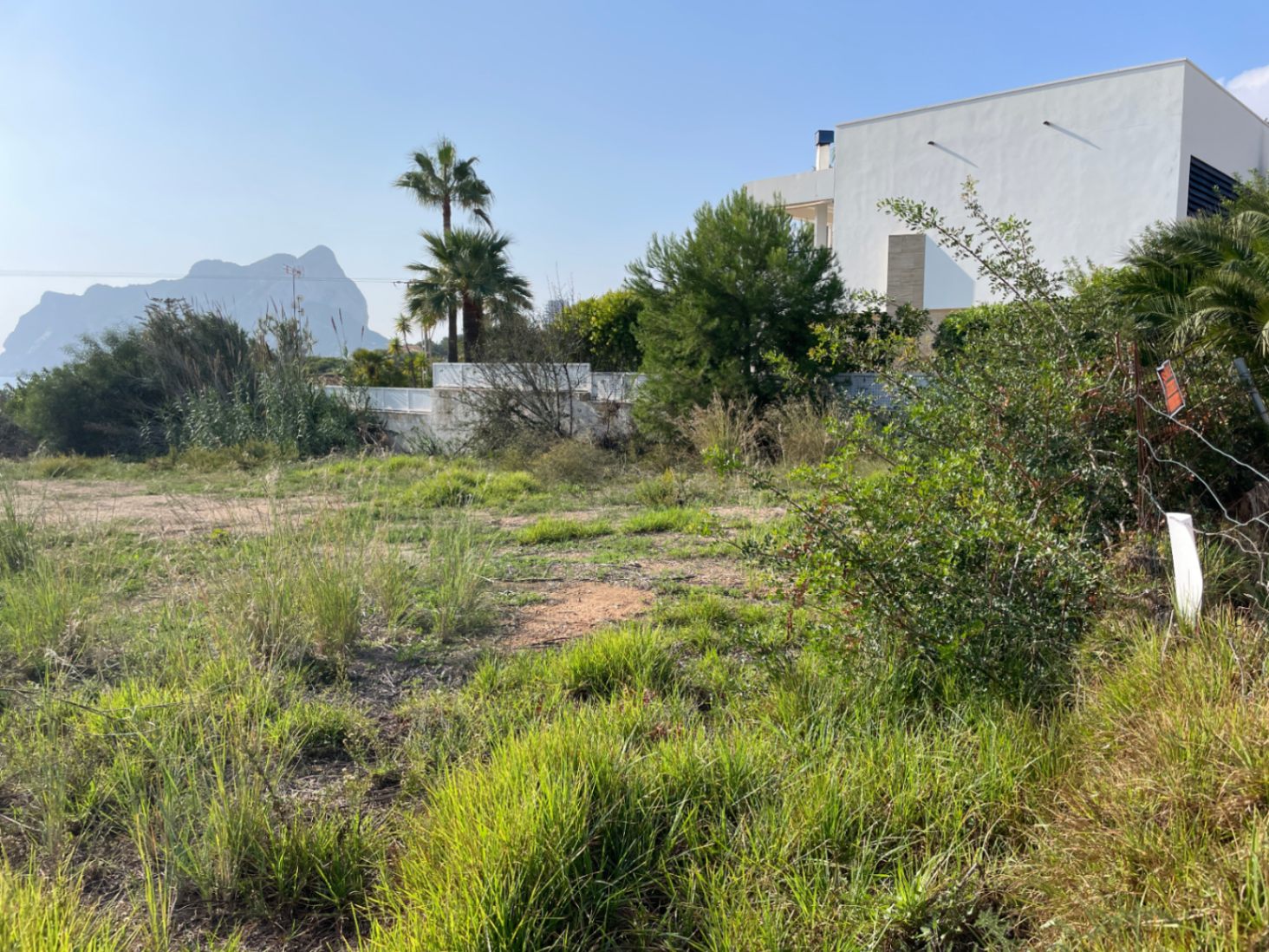 Parcela llana en Bassetes, Calpe, con vistas al Peñón de Ifach. El terreno cuenta con vegetación natural y linda con una propiedad de estilo moderno bajo un cielo despejado.