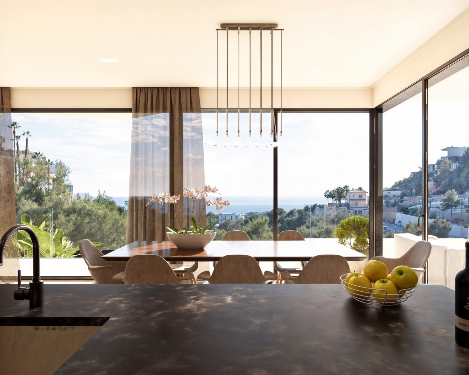 Comedor de concepto abierto con mesa de madera y vistas panorámicas al mar a través de grandes ventanales, integrado con una cocina de diseño de tonos oscuros.