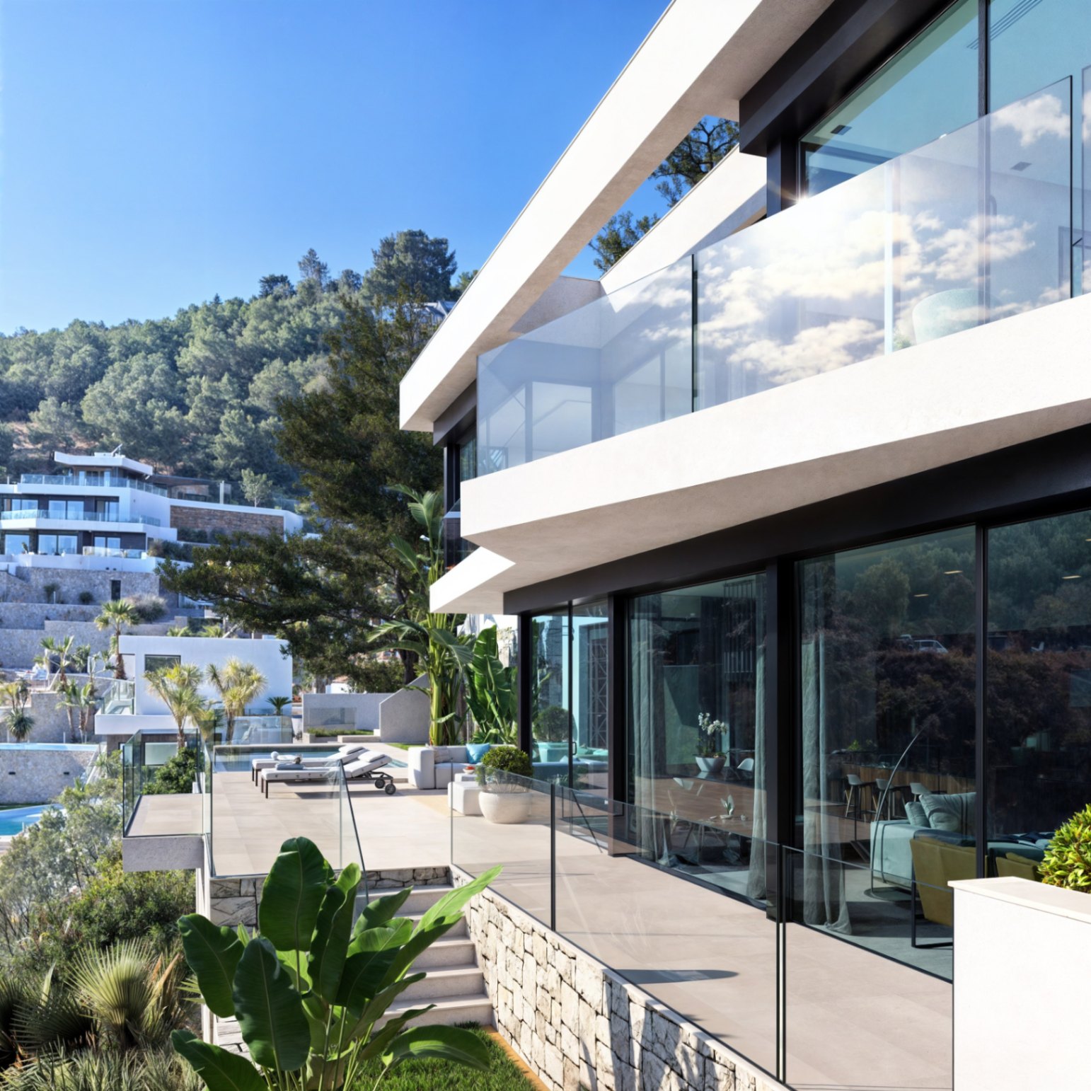 Fachada moderna de dos niveles con amplios ventanales, barandillas de cristal y terrazas de piedra natural integradas en un entorno de vegetación mediterránea.