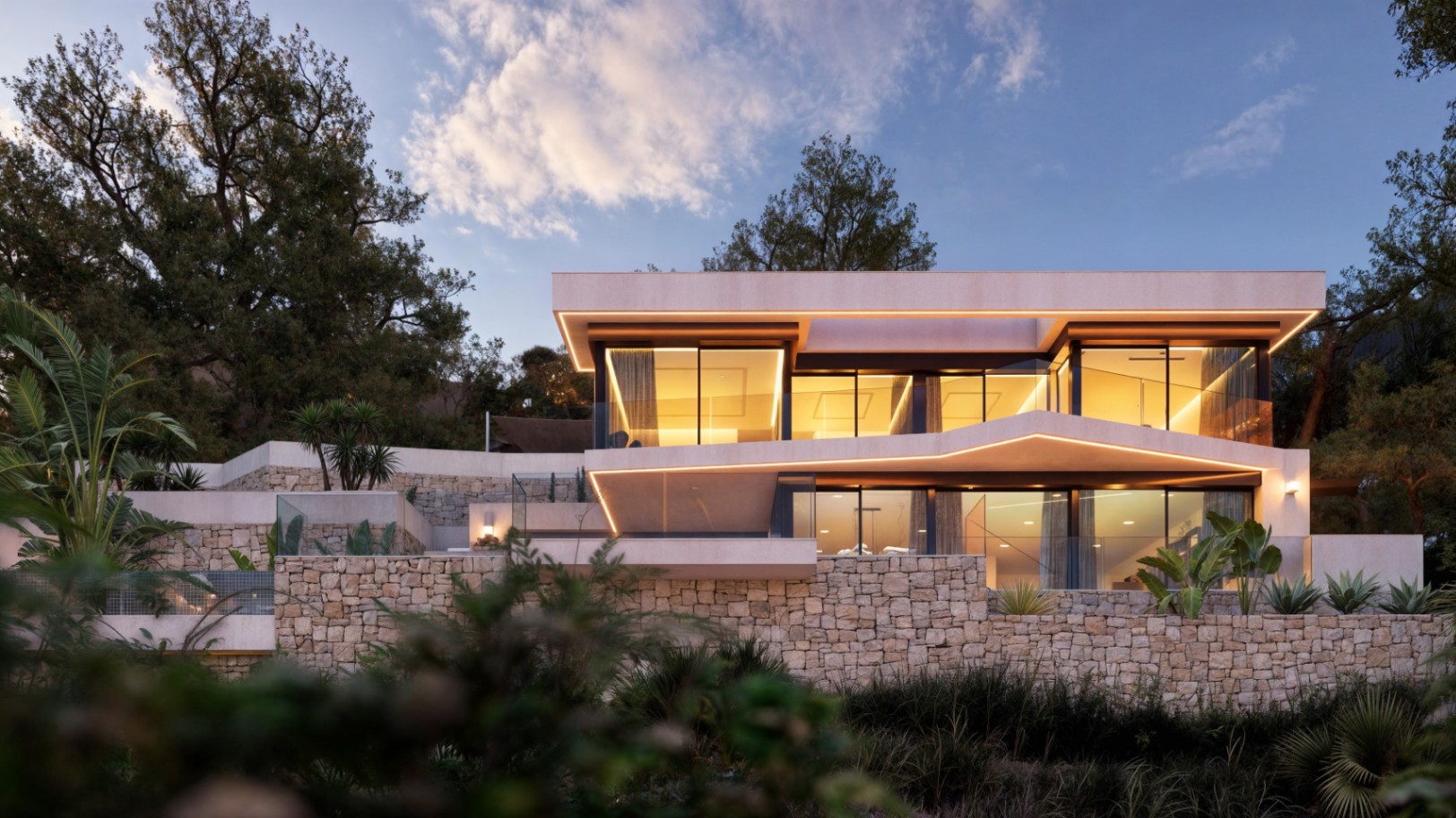 Fachada moderna de dos niveles con muros de piedra natural, iluminación LED integrada en voladizos y amplios ventanales que revelan interiores cálidos al atardecer.