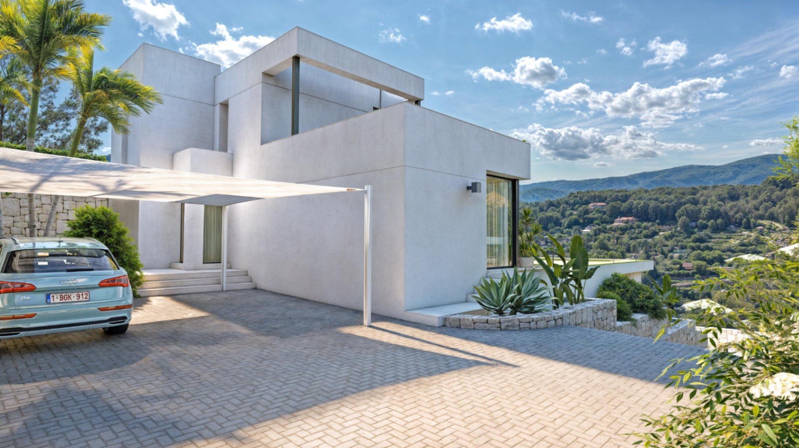 Fachada minimalista de líneas geométricas con zona de aparcamiento pavimentada, vela de sombra y muros de piedra natural frente a las vistas de la Sierra de Bernia.