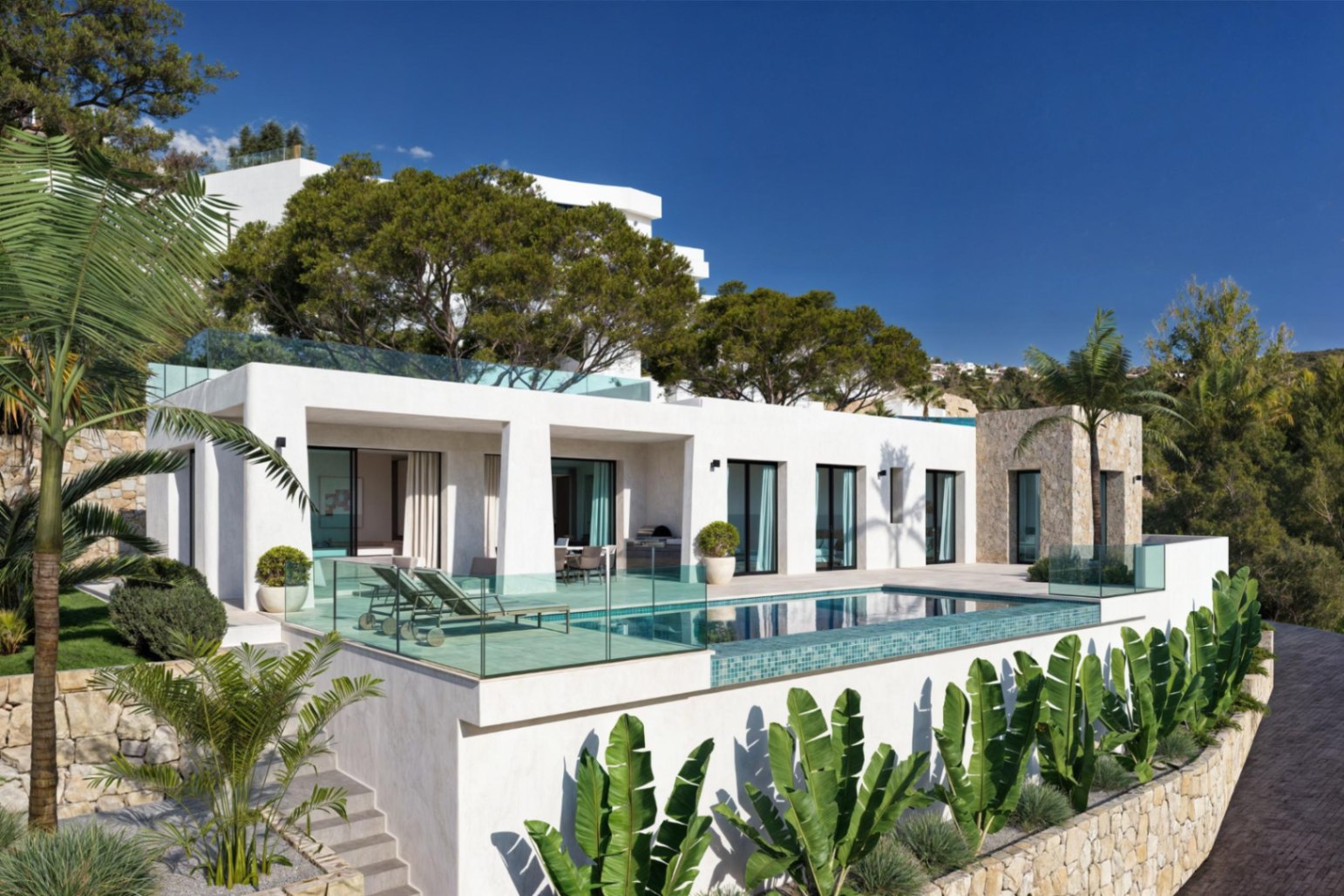 Vista exterior de la villa con piscina, terraza con tumbonas y jardín mediterráneo con palmeras.