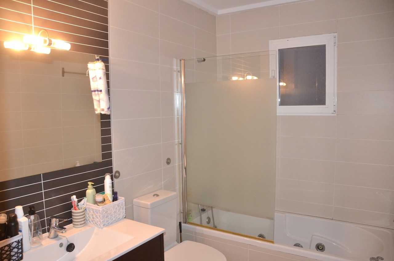 Baño moderno con lavabo integrado, mueble oscuro y bañera con mampara de cristal traslúcido. Paredes en tonos beige con un panel de contraste oscuro tras el espejo.
