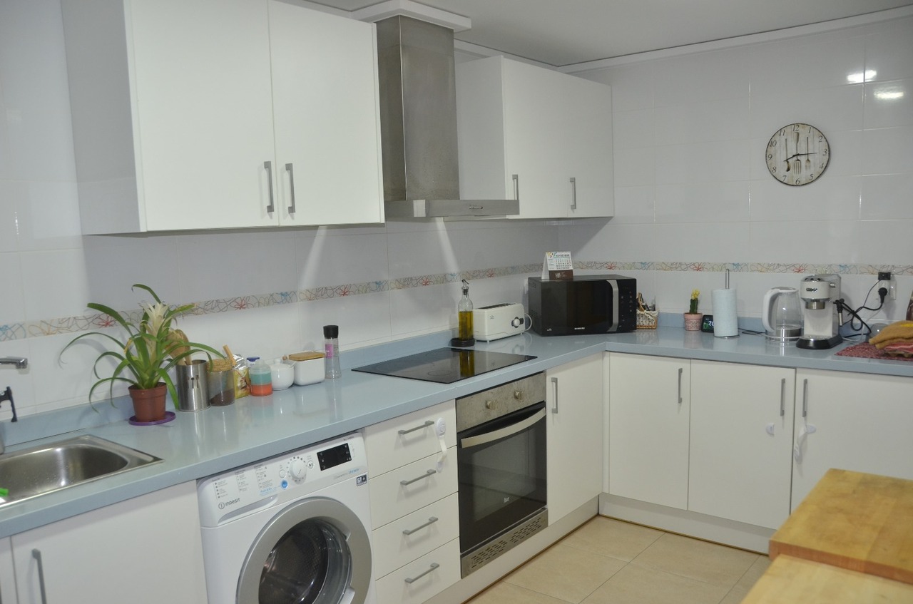 Cocina equipada con armarios blancos, encimera en tono azul claro, lavadora integrada, horno y placa vitrocerámica sobre paredes alicatadas.