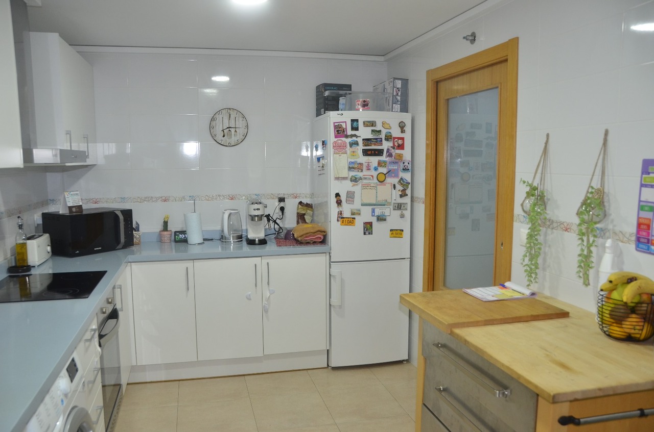 Cocina funcional con armarios blancos, encimera azul claro y electrodomésticos integrados. Incluye una isla de madera y paredes alicatadas con cenefa decorativa.