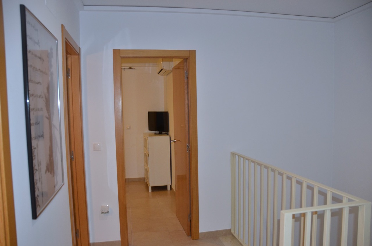 Distribuidor de planta superior con marcos de madera clara, barandilla metálica blanca y acceso a un dormitorio con cómoda blanca y televisión.