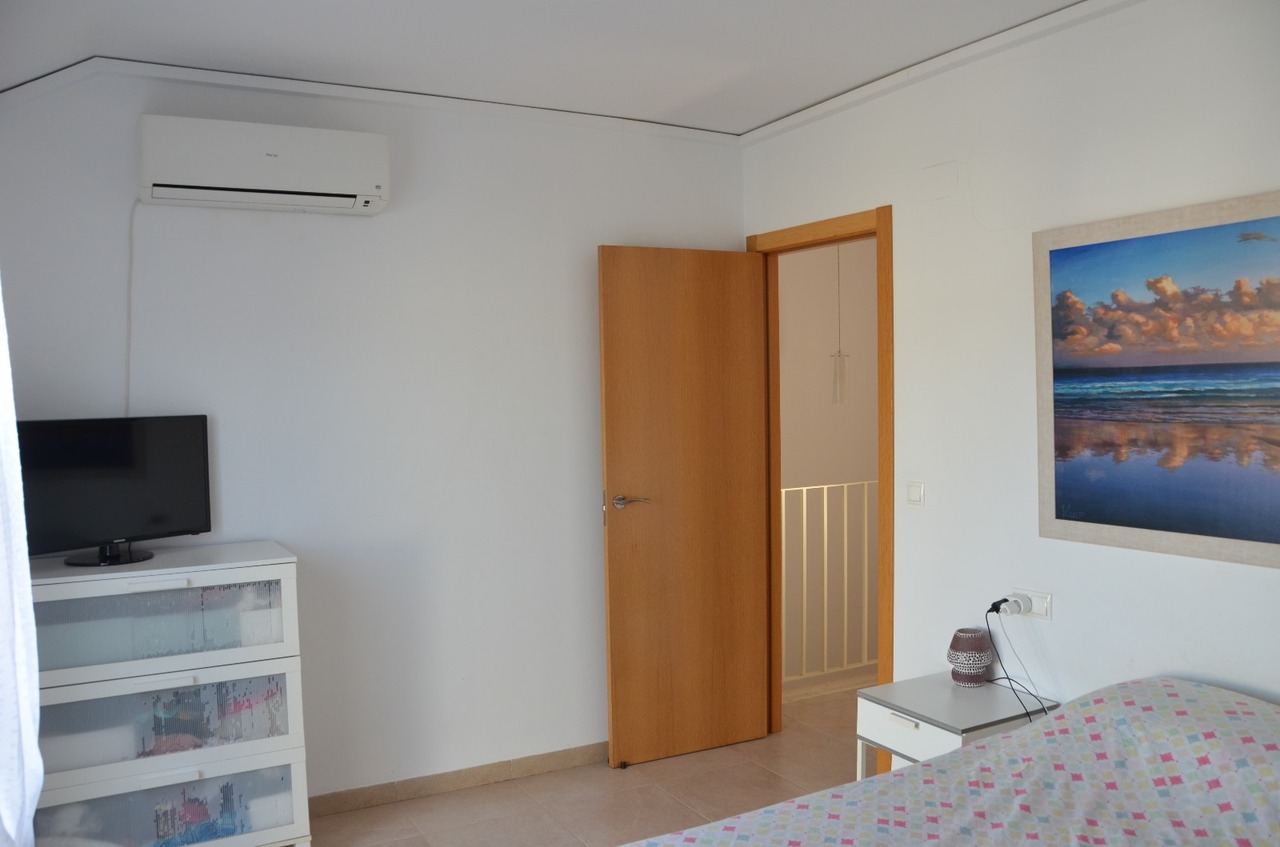 Dormitorio con aire acondicionado tipo split, cómoda blanca con televisor y puerta de madera hacia el pasillo. Presenta suelos cerámicos y decoración funcional.