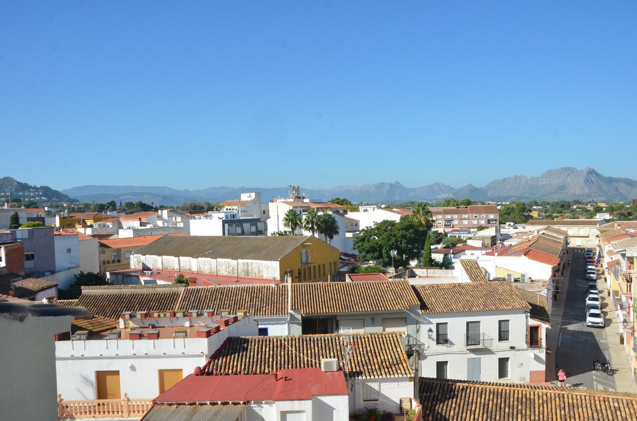 Vista panorámica elevada de los tejados de terracota y fachadas blancas del pueblo, con una cordillera montañosa al fondo bajo un cielo despejado.