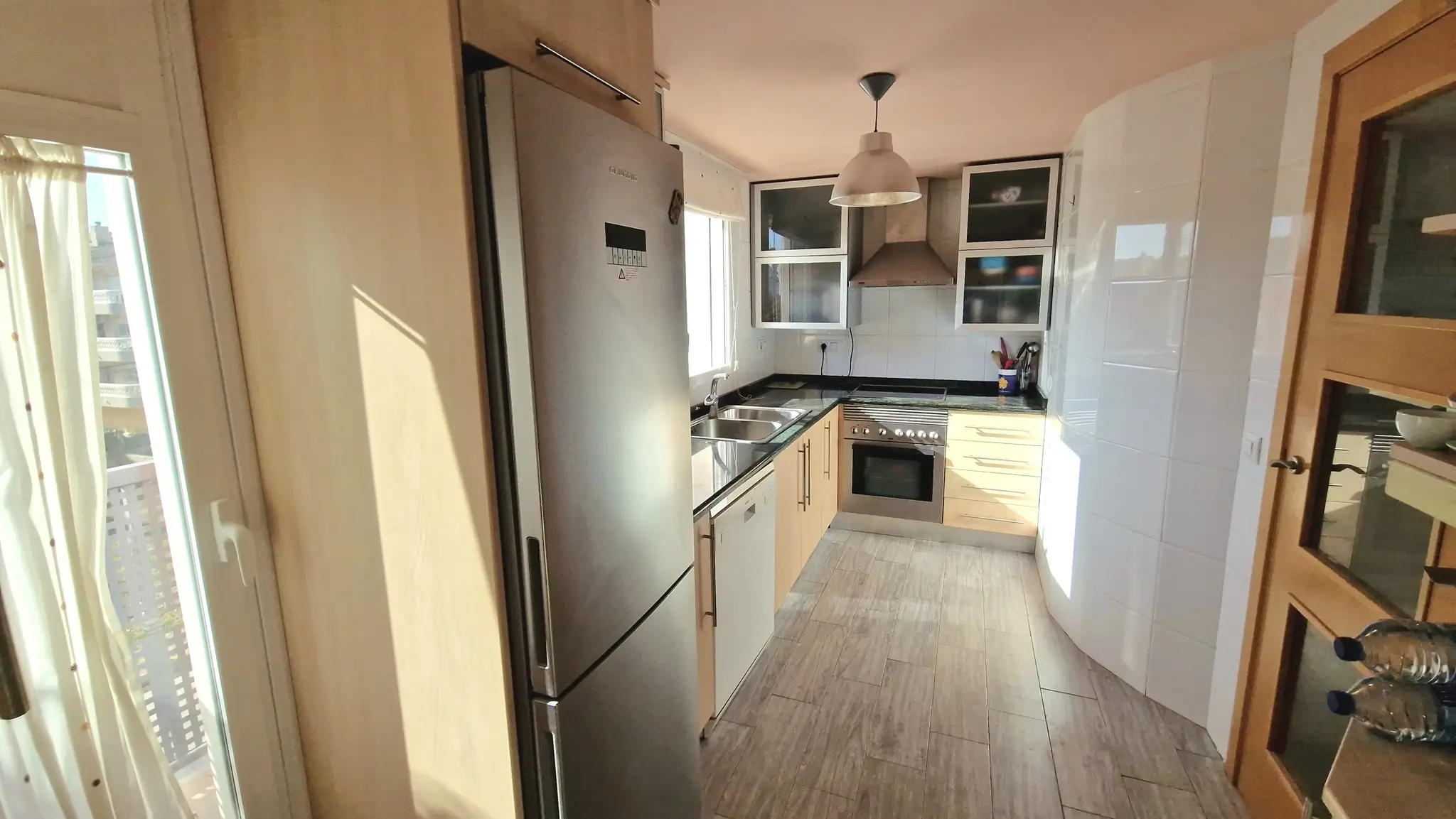 Cocina equipada con mobiliario de madera clara, encimeras de granito negro y electrodomésticos de acero inoxidable. Suelo de gres cerámico con efecto madera.
