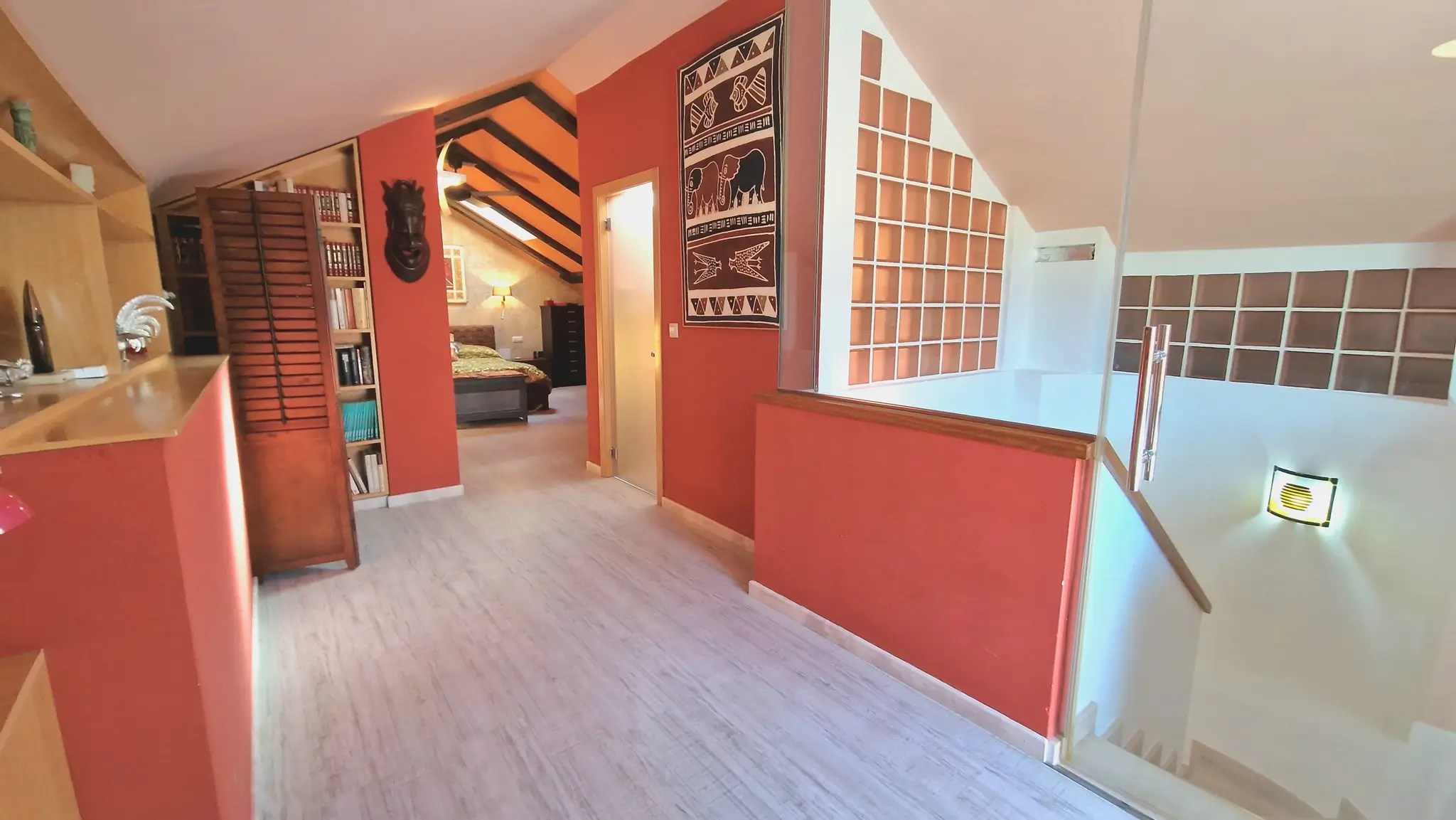 Distribuidor superior con paredes terracota, suelos de madera clara y barandilla de cristal. Al fondo, dormitorio abuhardillado con vigas de madera oscura y estanterías.